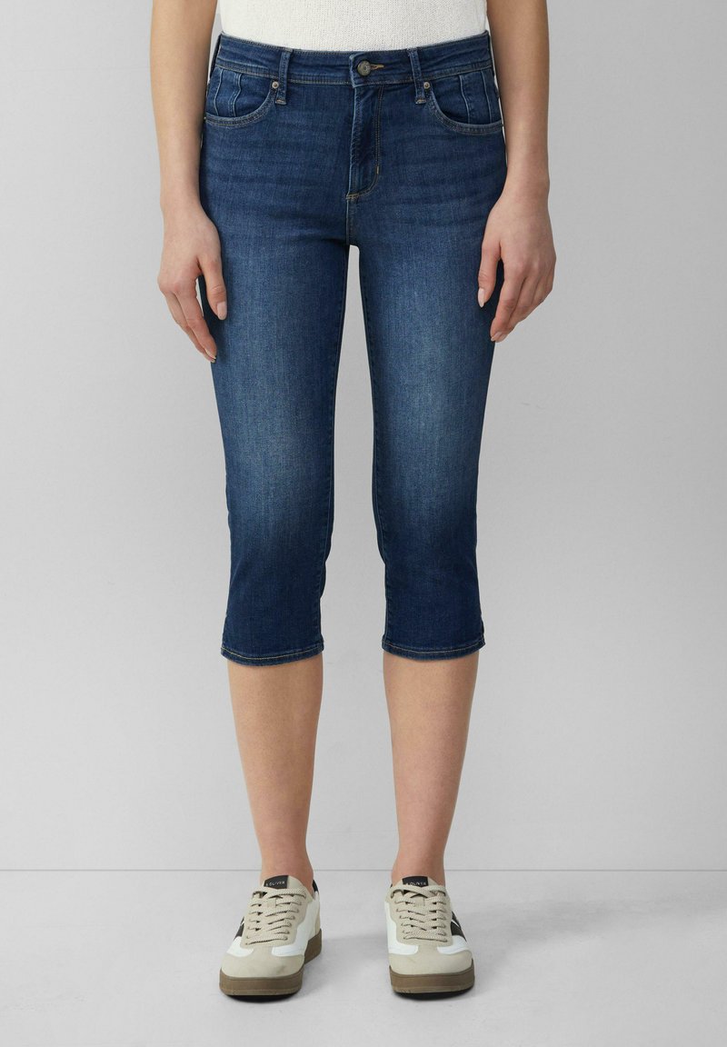 Tmavomodré denim capri nohavice so priliehavým strihom, stredne vysokým pásom, dvoma prednými vreckami a manžetami na koncoch. Nosené s bielymi teniskami.