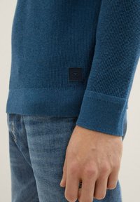 Suéter de punto texturizado azul con puños acanalados, que presenta una pequeña etiqueta negra en el dobladillo. Combinado con jeans de denim azul claros.