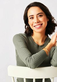 Modella che indossa un top a maniche lunghe verde oliva a coste, seduta su una sedia bianca, con un bracciale d'oro al polso e uno sfondo neutro.