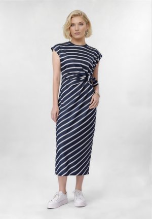 Szőke nő tengerészkék-fehér átlós csíkos midi ruhában, oldalt megkötve, fehér tornacipőben, karika fülbevalóval és karkötővel, fehér háttér előtt áll.