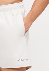 Balti šortai iš lengvos medžiagos, su elastinga juosmens juosta, šonine kišene ir juodu "Calvin Klein" logotipu ant pakraščio.