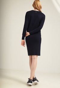 Robe longue à manches longues de couleur bleu marine avec une silhouette ajustée, texture tricotée, et un logo discret au dos, associée à des baskets à motifs noir et blanc.