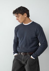 Marineblauwe trui met geribde hals en manchetten, boven een wit T-shirt. Gecombineerd met zwarte jeans en een bruine leren riem, met een zilveren gesp.