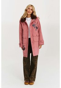 Ružový velúrový kabát s hnedým umelým kožuchom na golieri, prednými vreckami a vyšívanými vzormi medúzy a jablka. Skombinovaný s nohavicami s leopardím vzorom.
