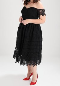 Černé krajkové šaty s odhalenými rameny, které mají vrstvené textury, složité vzory a zvlněný lem, doplněné červenými stiletto podpatky.