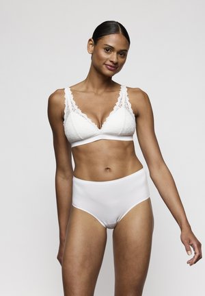 Mujer de cabello oscuro con un bralette de encaje blanco y ropa interior blanca de cintura alta, de pie contra un fondo claro y simple, sonriendo.