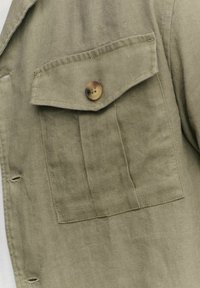 Olivengroene linnen blouse met een klepzak, voorzien van een ronde bruine knop en zichtbare stikseldetails. De stof heeft een zachte, gestructureerde afwerking.