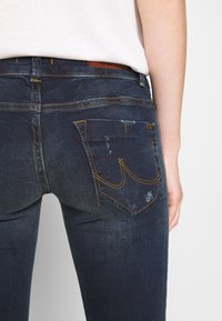 Jean en denim bleu foncé avec une coupe ajustée, présentant des surpiqûres jaunes, deux poches arrière avec des motifs de surpiqûres uniques et un léger effet usé.