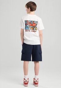 White t-shirt featuring a colorful Keith Haring graphic on the back, paired with navy cargo shorts and red sneakers.