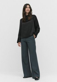 Sort satinbluse med cowl-neck og brede ærmer, parret med højtaljede, løstsiddende, mørkegrå bukser. Sorte slip-on sko.