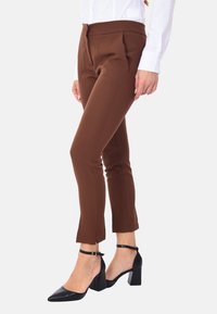 Pantaloni tapered marroni con pieghe frontali, realizzati in un tessuto liscio. Abbinati a scarpe nere con tacco a blocco, con cinturini alla caviglia.