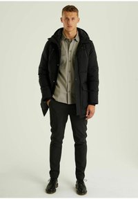 Schwarze Daunenjacke mit Kapuze, Reißverschluss und vorderen Taschen. Getragen über einem grauen Hemd mit Kragen und dunklen Hosen aus strukturiertem Material.