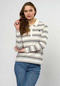 Vrouw met lang haar, gekleed in een crème polo met lange mouwen met zwarte en beige strepen en een blauwe jeans, staand tegen een effen lichte achtergrond.