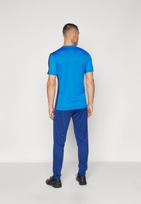 Blaues Sport-T-Shirt mit kurzen Ärmeln, kombiniert mit passenden blauen Hosen, beide aus glattem, leichtem Material. Schwarze Sportschuhe.