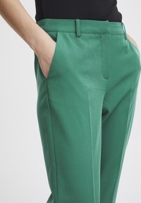Zelene krojene hlače glatke teksture, s bočnim džepovima, ravnim prednjim dijelom i prilagođenim strukom. Dizajnirane za elegantnu siluetu.
