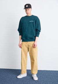 Felpa verde in cotone con collo rotondo, pantaloni beige morbidi e scarpe da ginnastica bianche, su uno sfondo di parete e moquette semplici.