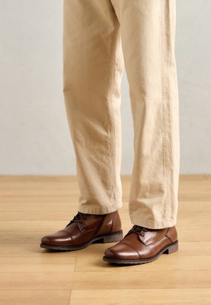 Chaussures habillées en cuir marron à lacets portées avec un pantalon beige à jambes droites sur un sol en bois clair.