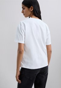 White cotton t-shirt with short sleeves, relaxed fit, and rear seam detail, paired with dark gray jeans against a neutral background.