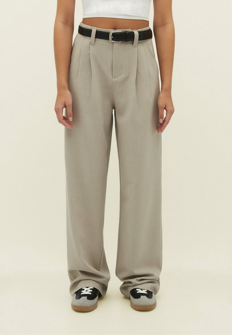 Personne portant un pantalon large beige avec des plis devant, une ceinture noire, un top court blanc et des baskets gris et noir avec des lacets blancs.