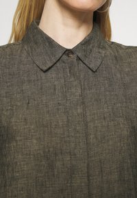 Primo piano di una persona che indossa una camicia di lino oliva scuro con colletto a punta e un unico bottone visibile fissato al collo.