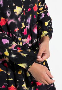Musta mekko, jossa on värikäs abstrakti kuvio, nappiyksityiskohtia, joustavat hihansuut ja kerätty vyötärö rentoa istuvuutta varten.
