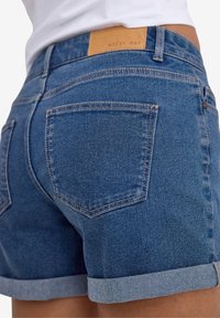 Denima īsās bikses vidēji zilā krāsā ar sarullētu apakšmalu, ar aizmugurējiem kabatām un gaiši brūnu uzlīmi "NOISY MAY" uz jostas.