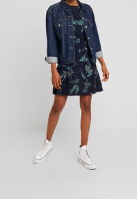 Jeansjacka med rullade ärmar över en marinblå blommig klänning med gröna och lila bladmönster. Bärs med vita högklackade sneakers.