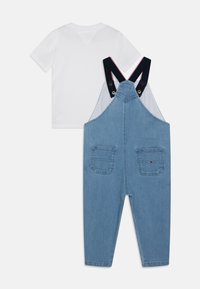 Denim tuinbroeken met verstelbare marineblauwe banden, voorzien van twee achterzakken en rode en witte accenten. Vergezeld van een plain wit T-shirt.