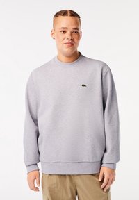 Jeune personne aux cheveux tressés portant un sweat-shirt gris clair avec un logo vert et un pantalon beige sur un fond blanc.