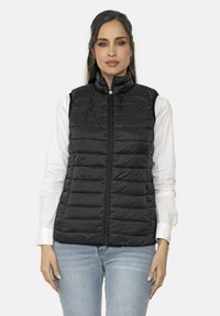 Chaleco acolchado negro con cuello alto, con costuras horizontales y una cremallera en la parte delantera. Lleva puesto encima de una camisa de manga larga blanca.