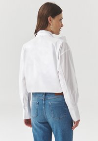 Camiseta blanca recortada con mangas largas y un cuello alto, combinada con pantalones vaqueros azules. Se pueden ver detalles de botones y una textura de tela suave.
