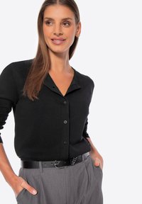 Camicetta nera con design a V, texture a coste e maniche lunghe, abbinata a pantaloni grigi e una cintura nera con borchie.