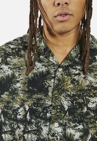 Korte mouw shirt met een groen en zwart grafisch patroon dat lijkt op bladeren. Gemaakt van een textuurstof met een puntige kraag en knoopsluiting aan de voorkant.