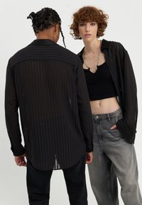 Svartstripet transparent skjorte med lange ermer og en lett buet hem, båret over en svart crop top og parret med grå denimjeans.