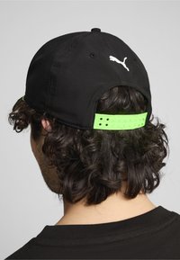 Juodas kepurė su kreivu snapu ir balta logotipu gale. Yra neon žalia reguliuojama juosta su skylutėmis dydžio reguliavimui.