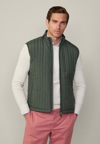 Grüne gesteppte Weste mit hohem Kragen, Reißverschlüssen und einer Seitentasche; über einem weißen Pullover getragen. Rosa Hose ergänzt das Outfit.