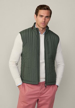Gilet matelassé vert avec un col montant, des élastiques et une poche latérale ; porté par-dessus un pull blanc. Un pantalon rose complète la tenue.