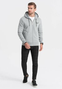 Grauer Reißverschluss-Hoodie mit gefütterter Kapuze, Fronttaschen und gestreiften Ärmeln, kombiniert mit karierten schwarzen Hosen und schwarzen Turnschuhen.