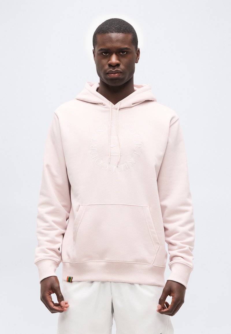 Homme portant un sweat à capuche rose pâle avec une poche avant et un short blanc, debout devant un fond gris clair uni.