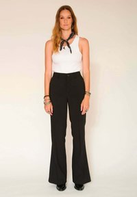 Pantalons évasés noirs taille haute avec une fermeture à bouton, associés à un débardeur blanc et une écharpe noire à motif. Bijoux en argent en accent.