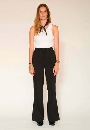 Pantalons évasés noirs taille haute avec une fermeture à bouton, associés à un débardeur blanc et une écharpe noire à motif. Bijoux en argent en accent.