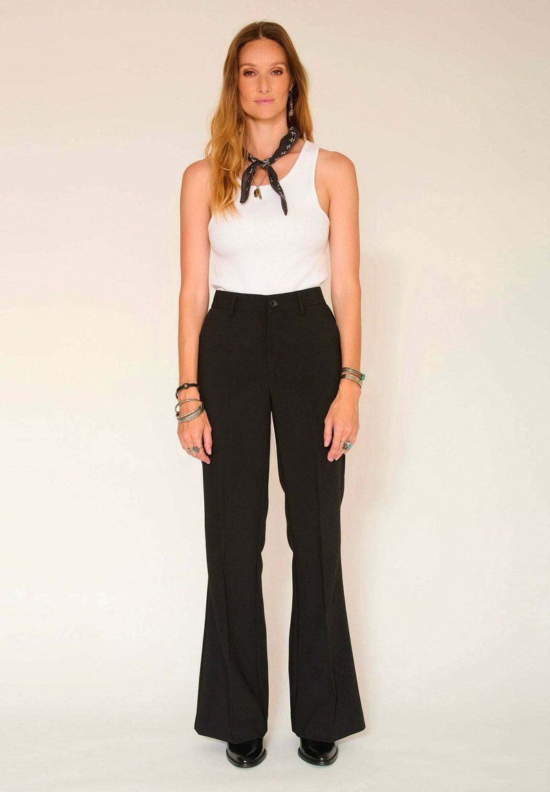 Pantalons évasés noirs taille haute avec une fermeture à bouton, associés à un débardeur blanc et une écharpe noire à motif. Bijoux en argent en accent.