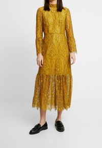 Vestido midi de encaje en amarillo mostaza con mangas largas, cuello alto y bajo con volantes. Presenta patrones florales intrincados y textura translúcida.