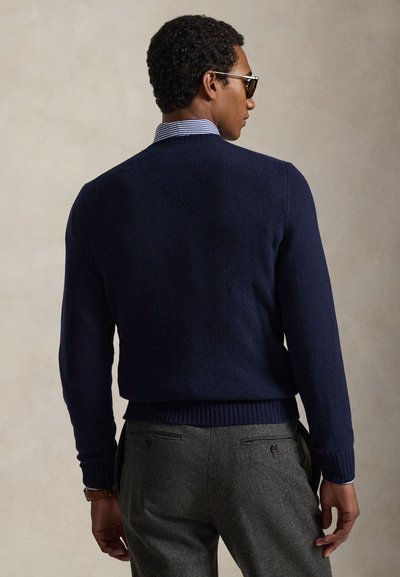 Man die een marineblauwe trui draagt over een gestreept overhemd en grijze broek, van achteren gezien, met een zonnebril en een horloge tegen een effen achtergrond.