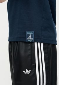 Temno modra Adidas majica z našitim značkom in črne Adidas trenirke s belimi črtami in trefoil logotipom na strani.