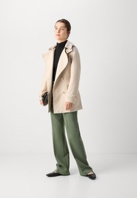 Beige trenchcoat met een brede kraag en dubbele knopen, gecombineerd met groene wijde broek en zwarte platte schoenen.