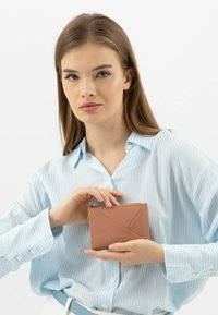 Portafoglio in pelle rosa compatto con design geometrico, dotato di una zip dorata e una texture liscia, tenuto da una persona con una camicia a righe blu.