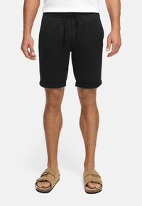 Schwarze Baumwollshorts mit elastischem Bund und Kordelzug, mit umgeschlagenen Säumen und Seitentaschen. Kombiniert mit braunen Slipper-Sandalen.