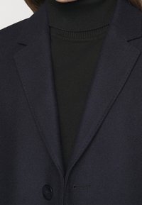 Blazer marine en tissu lisse, avec des revers à encoches et une fermeture à un bouton, superposé sur un pull col roulé noir à côtes.