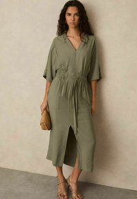 Olijfgroen midi-jurk met een V-hals, korte mouwen, elastische tailleband, voorknip en twee zakken. Gecombineerd met een gevlochten clutch en sandalen.